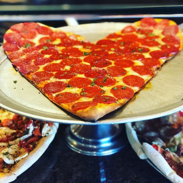 Heart-shaped pepperoni pizza on a metal stand, with other pizzas blurred in the background.