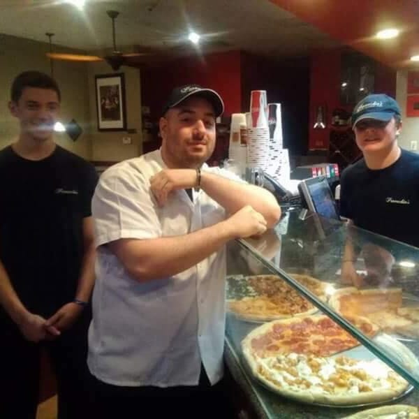 Three Pennachio's employees stand behind a counter displaying several fresh pizzas.