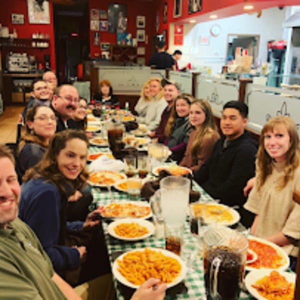 A large group of people enjoying pizza and pasta at Pennachio's, seated at a long table.