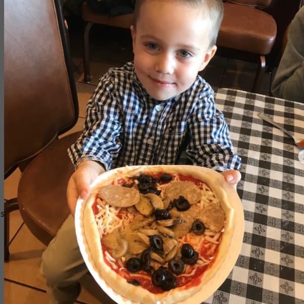 A young boy in a plaid shirt proudly holds up an uncooked Pennachio's pizza with toppings.