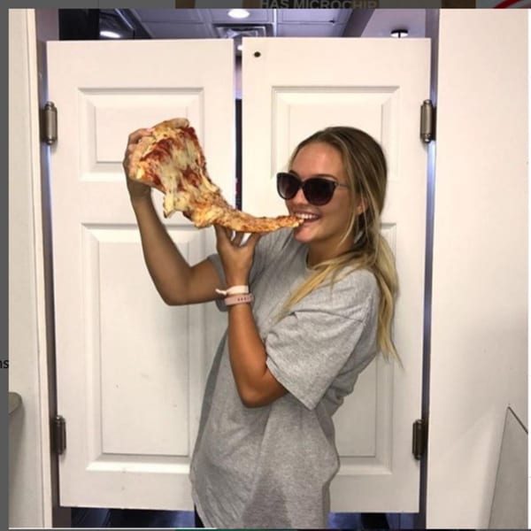 A young woman in sunglasses smiles while taking a bite from a large slice of Pennachio's pizza.