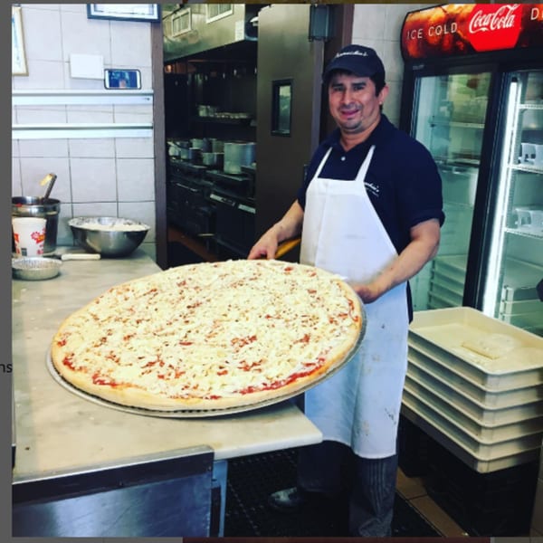 A Pennachio's chef holds a large, uncooked cheese pizza in a commercial kitchen.
